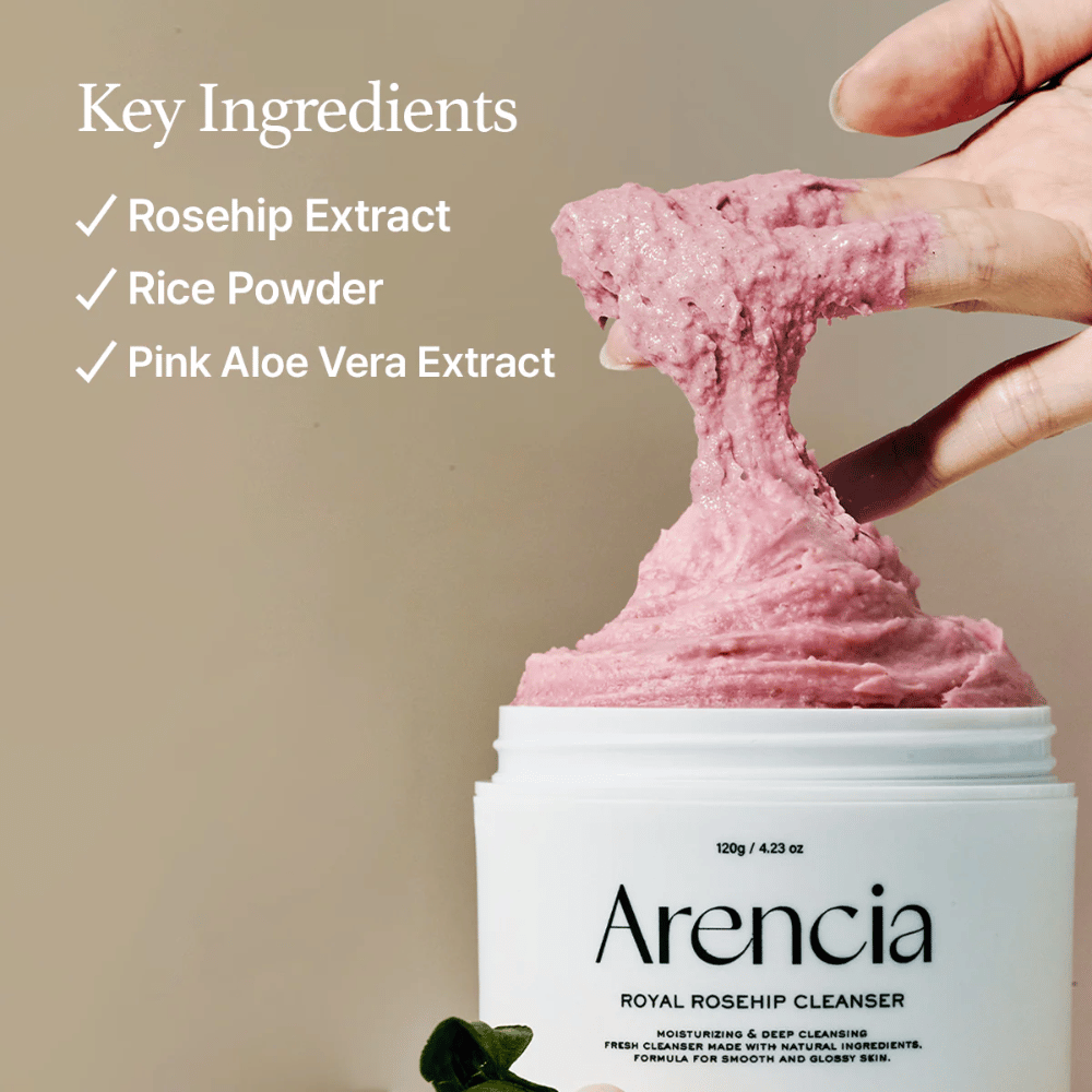 Una mano levanta la textura de limpieza rosa y esponjosa del tarro Arencia Royal Rosehip Cleanser, con la lista de ingredientes a la izquierda: extracto de rosa mosqueta, polvo de arroz y extracto de aloe vera rosa.