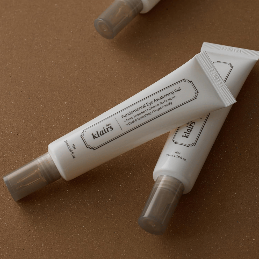 Imagen de estilo de vida con dos tubos de Klairs Fundamental Eye Awakening Gel colocados sobre un fondo marrón cálido. La composición resalta el diseño elegante del producto y su sensación premium, al mismo tiempo que pone el foco en el cuidado de los ojos y la hidratación.