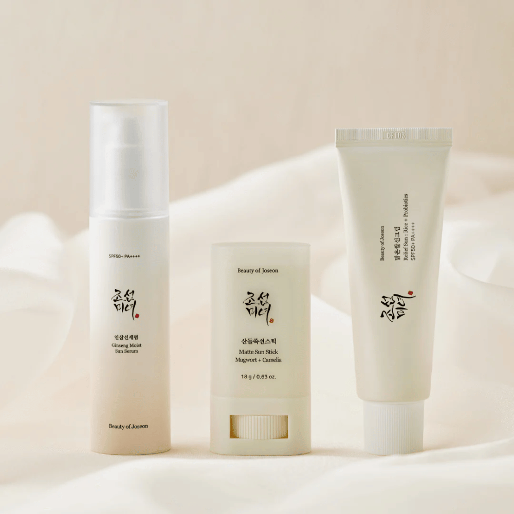 Tres productos de protección solar de Beauty of Joseon – Ginseng Moist Sun Serum, Matte Sun Stick y Relief Sun Rice + Probiotics – están alineados sobre una tela color crema con luz suave.