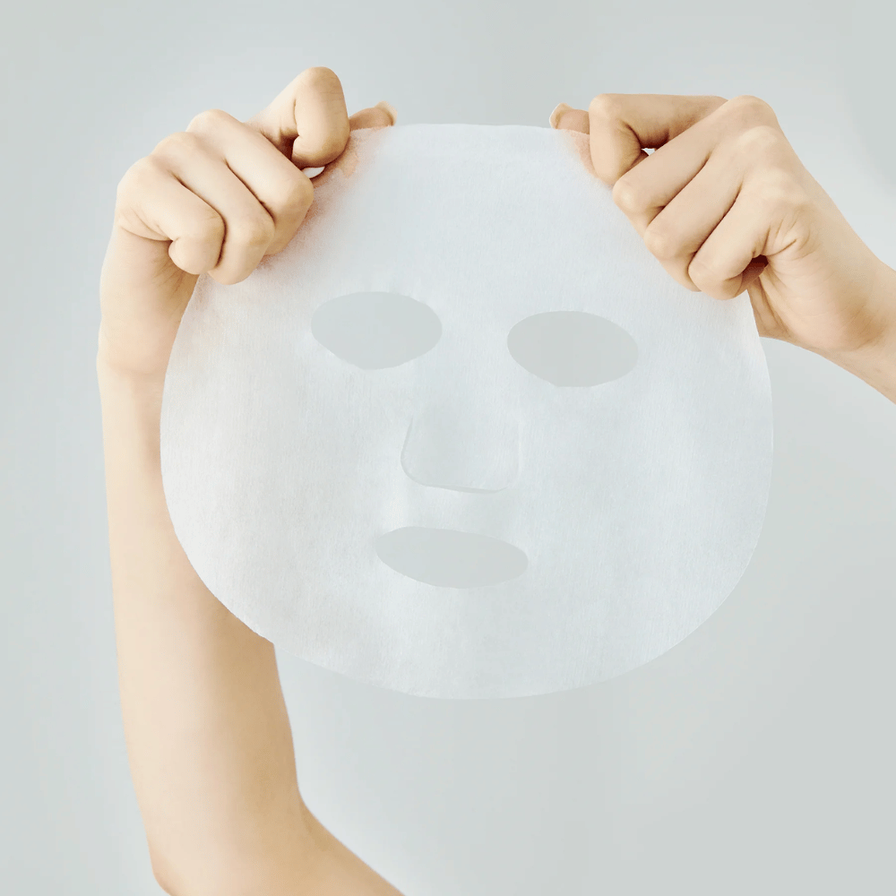Dos manos sostienen una mascarilla de hoja blanca contra un fondo gris claro, con recortes visibles para los ojos, nariz y boca.