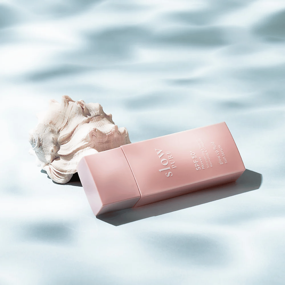 Slow Pure Glow Up Fluid Sun Serum junto a conchas marinas y efecto de agua, destaca protección solar y brillo.