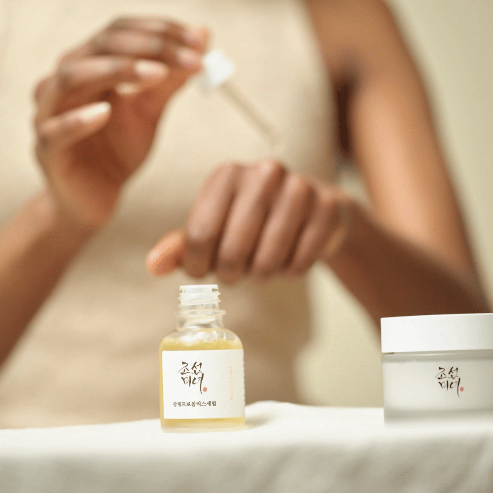 Persona aplicando Beauty of Joseon Glow Serum con pipeta de botella de vidrio, con un frasco de la misma serie al lado en la mesa.