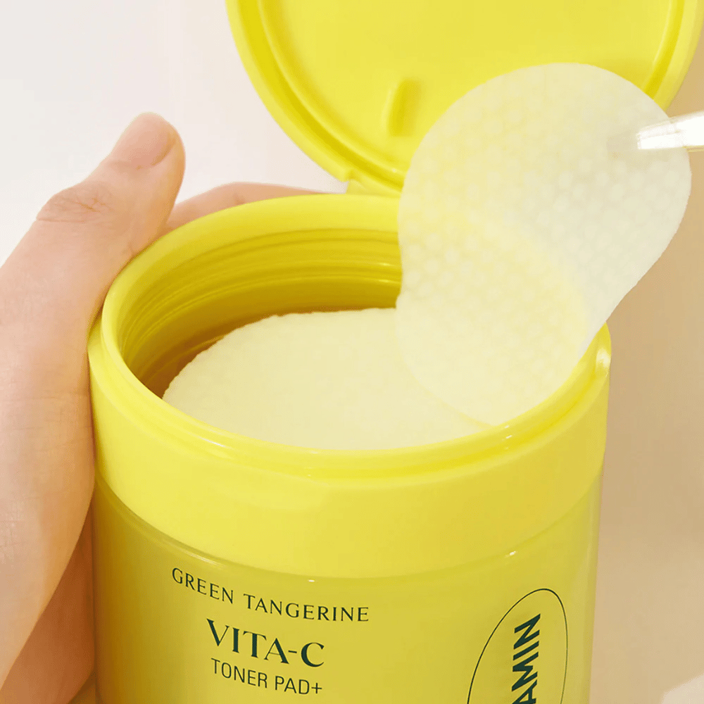 Una mano usa unas pinzas de plástico para recoger una almohadilla de tónico con textura de un contenedor amarillo claro marcado "GREEN TANGERINE VITA-C TONER PAD+" de la marca "gooda".