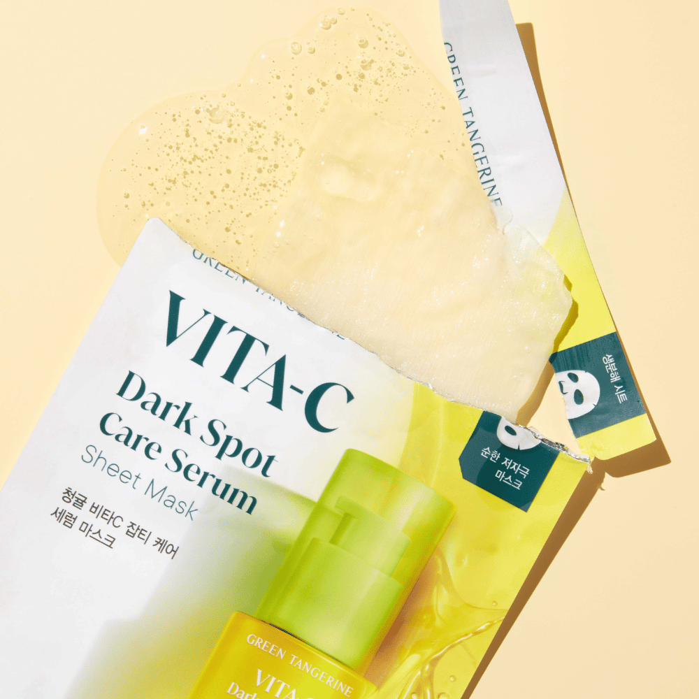 Envase abierto de VITA-C Dark Spot Care Serum Sheet mask con una mascarilla facial encima y una botella de suero verde, todo sobre un fondo amarillo.