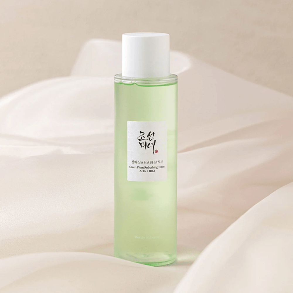 Una botella de "Green Plum Refreshing Toner" AHA + BHA está sobre un fondo suave y claro. El producto tiene un líquido verde que refleja frescura y naturalidad. La etiqueta es blanca con detalles en negro y rojo, lo que le da un aspecto elegante y limpio.