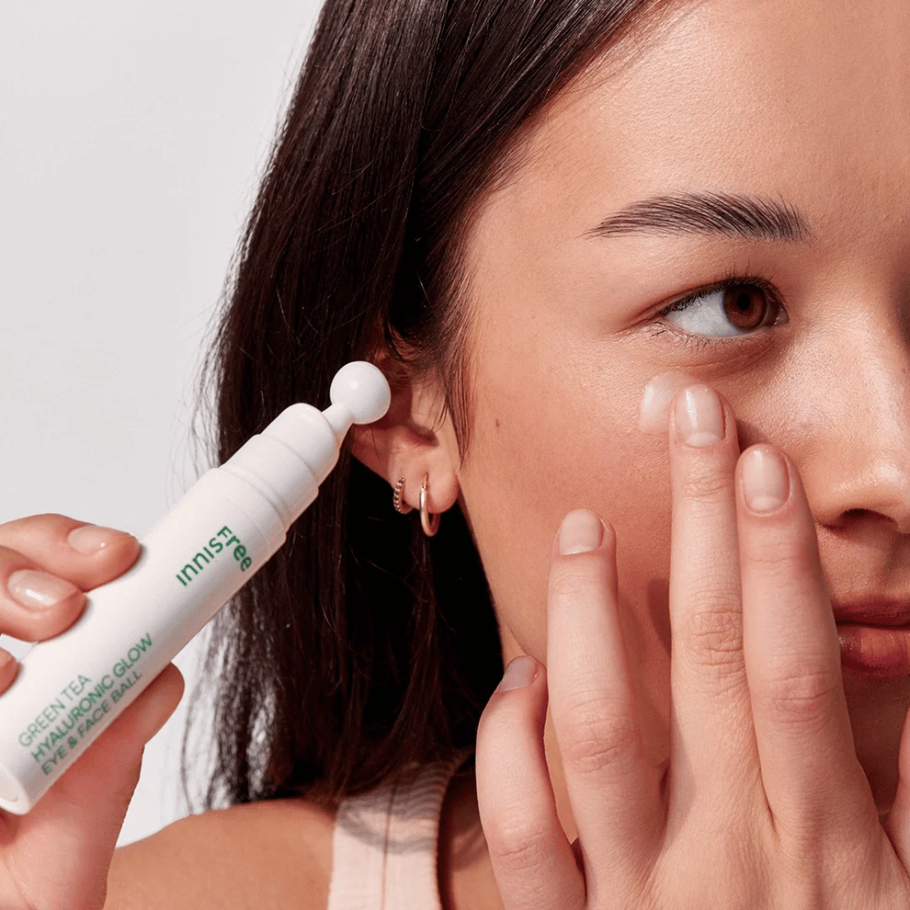 Una persona aplica Innisfree Green Tea Hyaluronic Glow Eye & Face Ball en su piel debajo de los ojos. El producto tiene una parte aplicadora redonda y blanca y está en una botella blanca con texto verde. El rostro del usuario se ve parcialmente y se enfoca en la aplicación del producto.