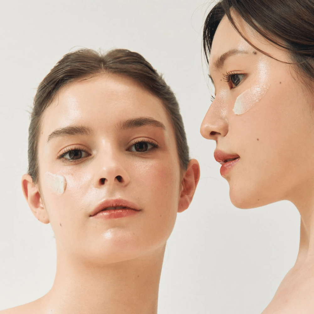 Dos mujeres con mascarillas aplicadas en partes de sus rostros. Una de las mujeres tiene la mascarilla en la mejilla y la nariz, mientras que la otra la tiene debajo del ojo y en la mejilla. Llevan maquillaje natural y se ven frescas, lo que resalta el efecto hidratante y luminoso de las mascarillas.
