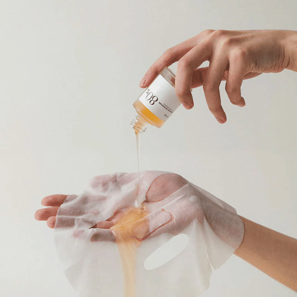 Una mano vierte un líquido espeso y dorado de una pequeña botella marcada '108' sobre una máscara facial transparente sostenida en otra mano. El fondo es neutral y uniforme para resaltar el producto.