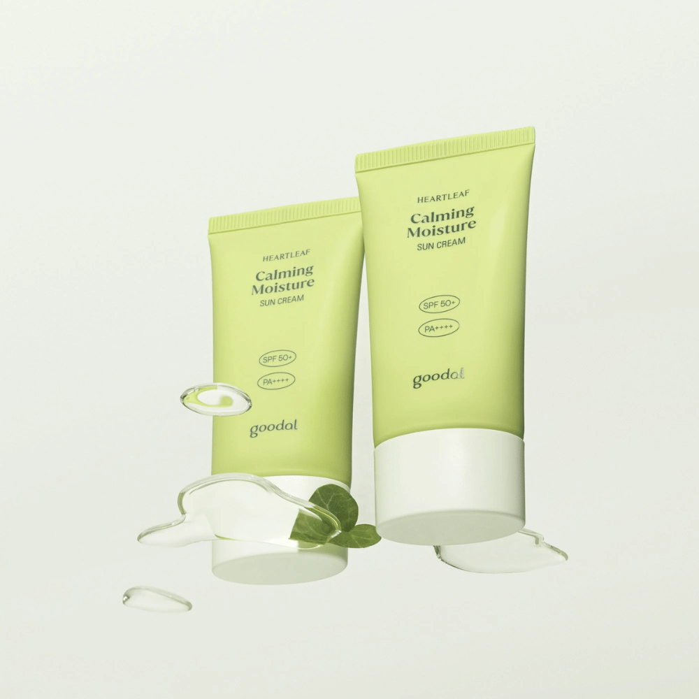 Dos tubos de Goodal Heartleaf Calming Moisture Sun Cream SPF50+ PA++++ rodeados de hojas de plantas y gotas de suero sobre un fondo claro.