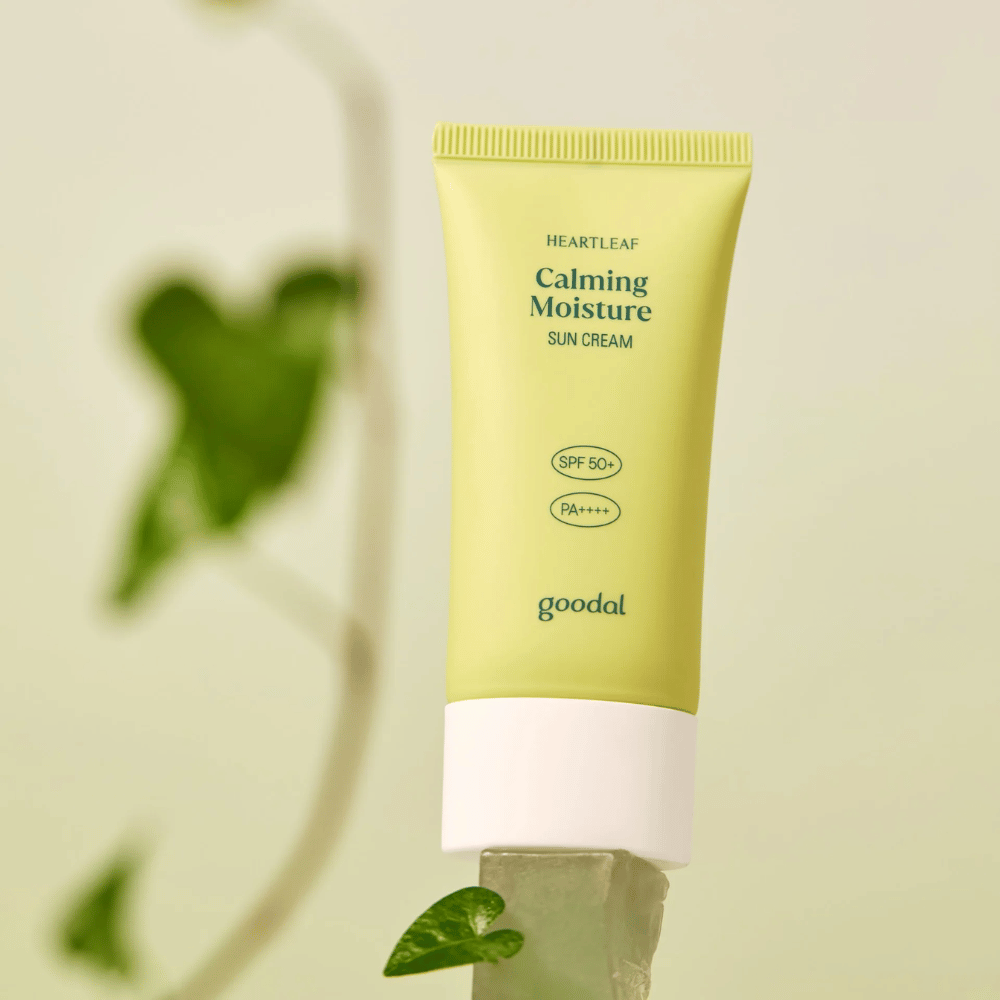 Goodal Heartleaf Calming Moisture Sun Cream SPF50+ PA++++ colocada sobre una lámina de vidrio con una planta verde en el fondo - un calmante protector solar para piel sensible.