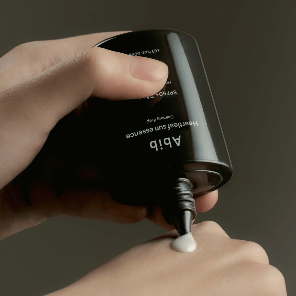 Mano exprimiendo Abib Heartleaf Sun Essence Calming Drop en el dorso de otra mano, con fondo oscuro y clara visualización de la consistencia del producto.