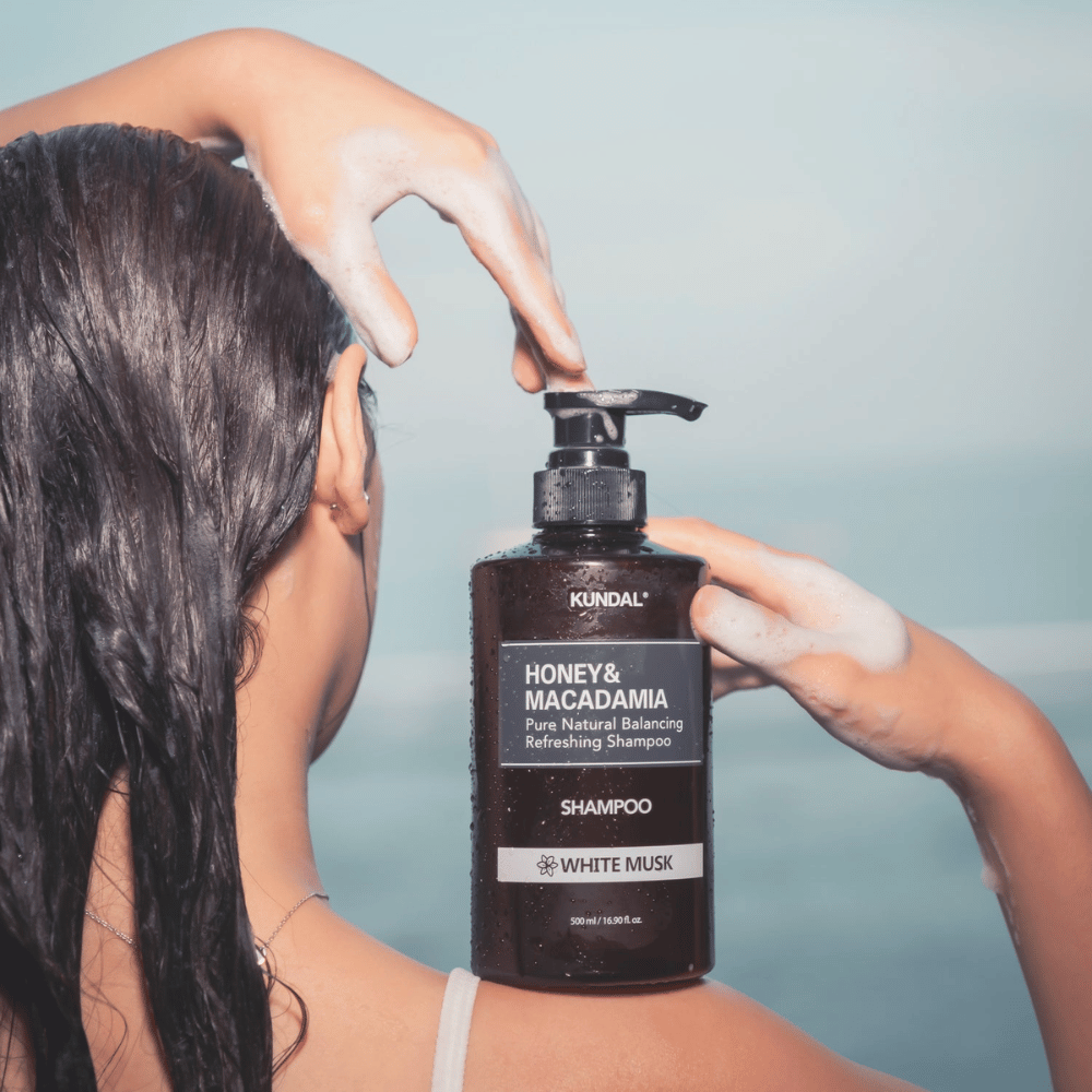 Mujer con el cabello mojado aplica espuma y sostiene KUNDAL Honey & Macadamia Nature Shampoo White Musk contra un fondo de mar.