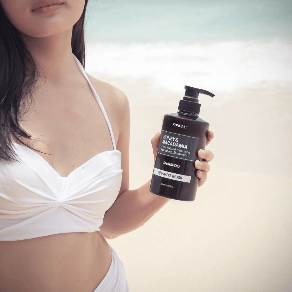 Mujer en bikini blanco sostiene KUNDAL Honey & Macadamia Nature Shampoo White Musk en una playa con el mar de fondo.