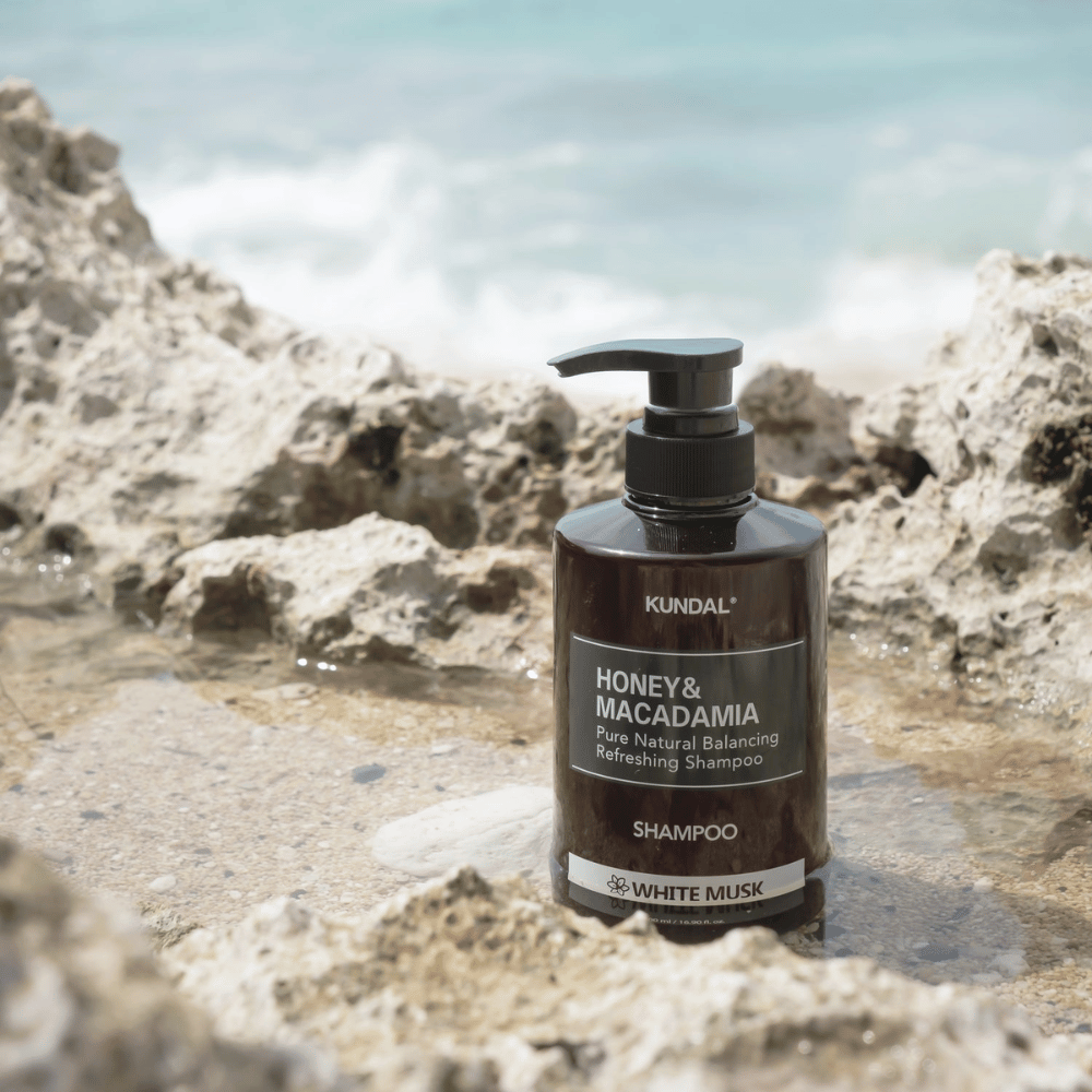 KUNDAL Honey & Macadamia Nature Shampoo White Musk colocada en las rocas junto al mar, un producto natural para el cuidado del cabello con un aroma fresco.
