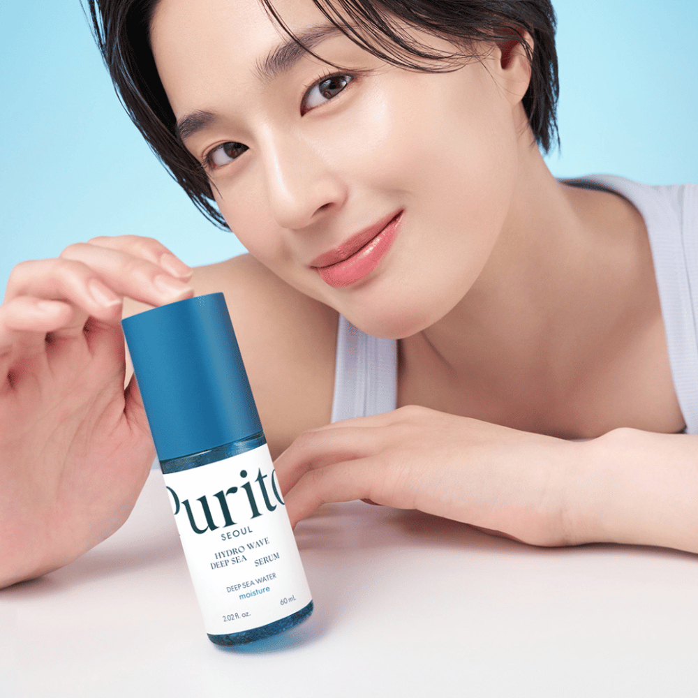 Una mujer con el pelo corto sostiene un suero azul de Purito, Hydro Wave Deep Sea Serum. Su piel se ve radiante e hidratada, lo que resalta el producto en foco. El fondo es azul claro, lo que proporciona una sensación fresca y limpia. El producto está enfocado en el cuidado de la piel.