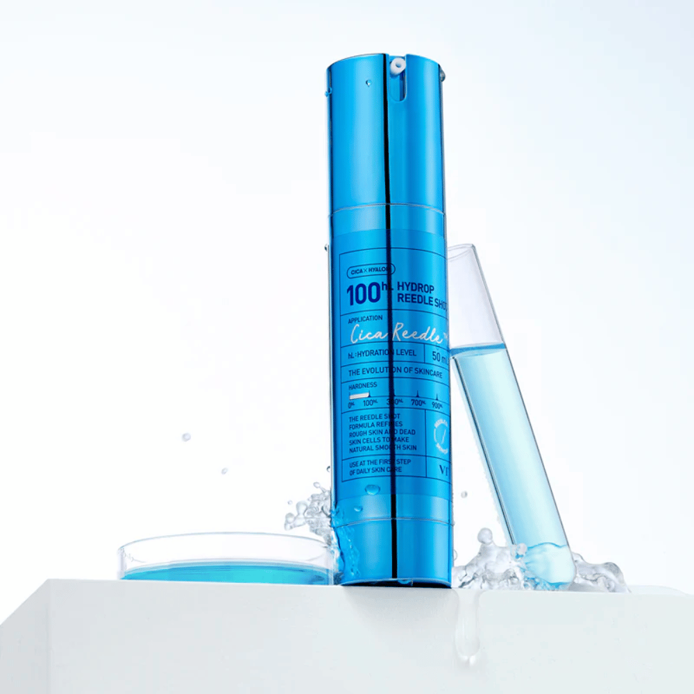 Botella azul VT Cica Hyalon Hydrop Reedle Shot 100hL de pie sobre superficie blanca con salpicaduras de agua, junto a un tubo de ensayo lleno de líquido azul y una tapa redonda con el mismo líquido.