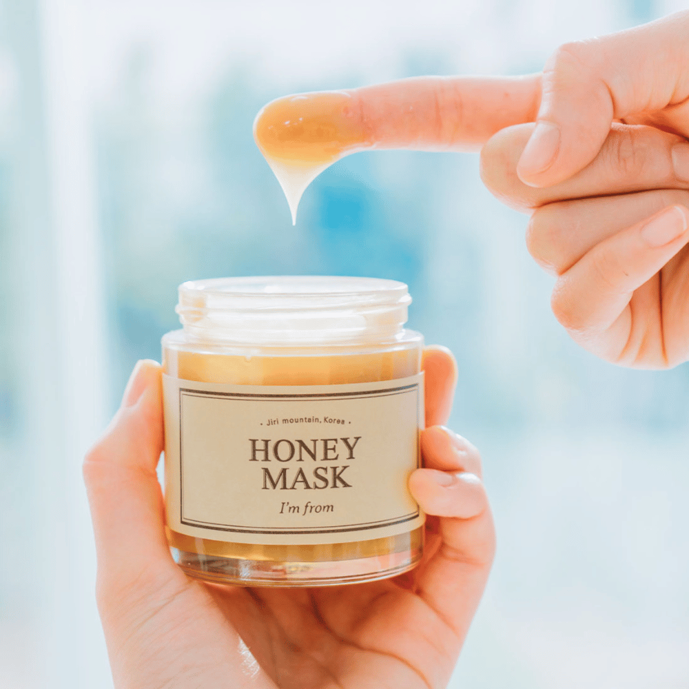 Una mano sostiene un tarro abierto de "I'm from Honey Mask", y un dedo se sumerge en la máscara dorada sobre un fondo claro.