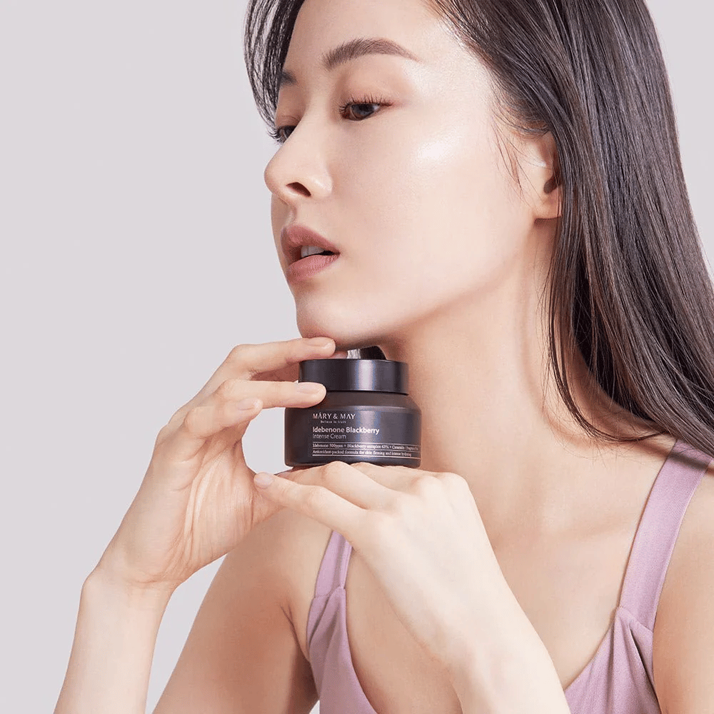 Una mujer sostiene un frasco con MARY & MAY Idebenone Blackberry Intense Cream. Ella tiene una piel radiante y sonríe ligeramente, vestida con un top sin mangas. El producto es una crema intensa con idebenona y complejo de mora destinada a la firmeza e hidratación de la piel.
