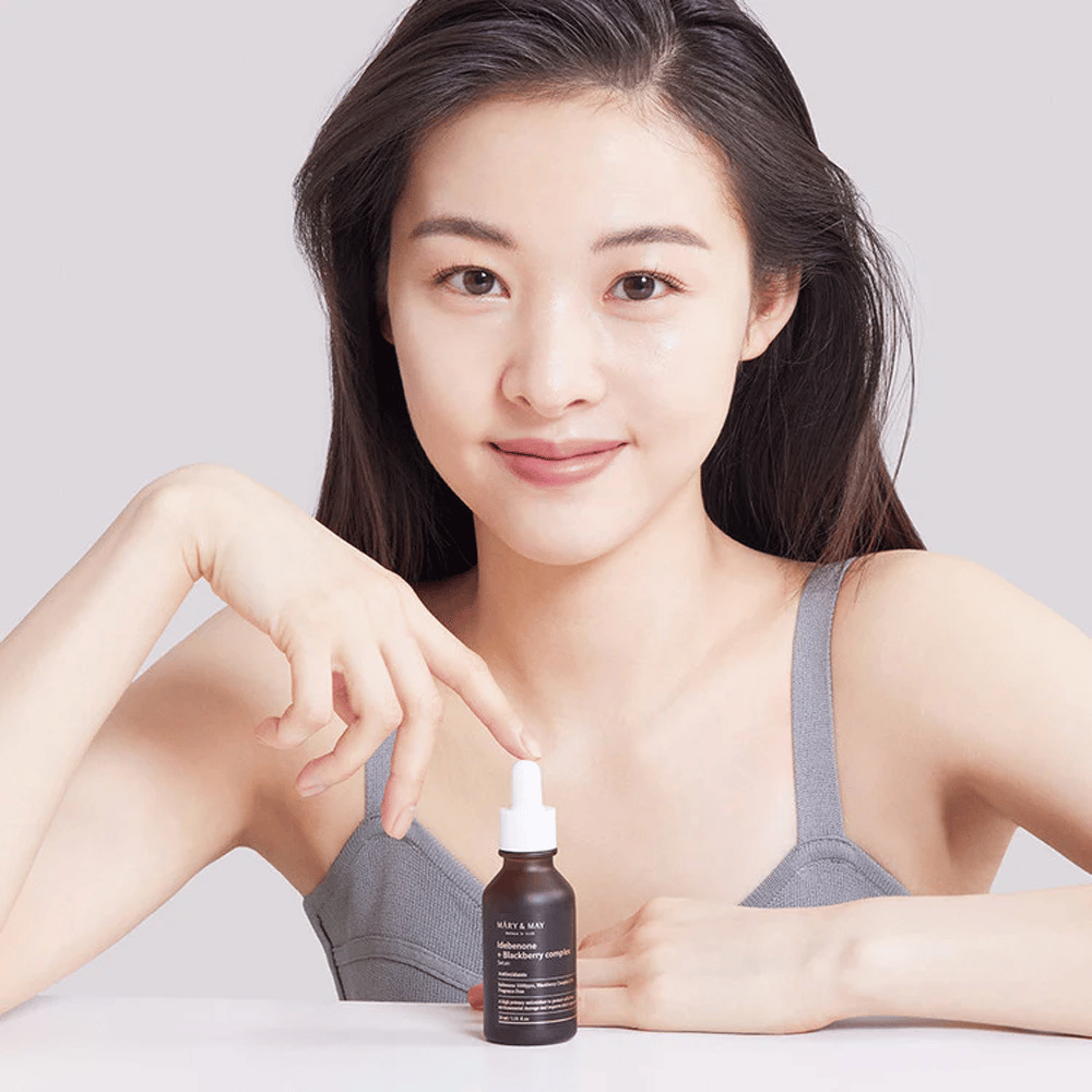 Una mujer con cabello largo y oscuro sostiene MARY & MAY Idebenone + Blackberry Complex Serum, una botella de vidrio oscuro con pipeta. Ella tiene una sonrisa alegre y señala la botella con su otra mano. La mujer está vestida con una blusa gris sin mangas y parece estar en un entorno luminoso y sencillo, lo que resalta el enfoque del producto en el cuidado de la piel y la belleza.