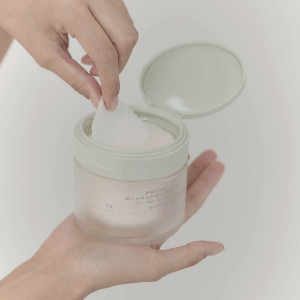 Mano sacando una almohadilla de tónico del tarro Celimax Ji Woo Gae Cica BHA Blemish Toner Pad, perfecta para piel impura.
