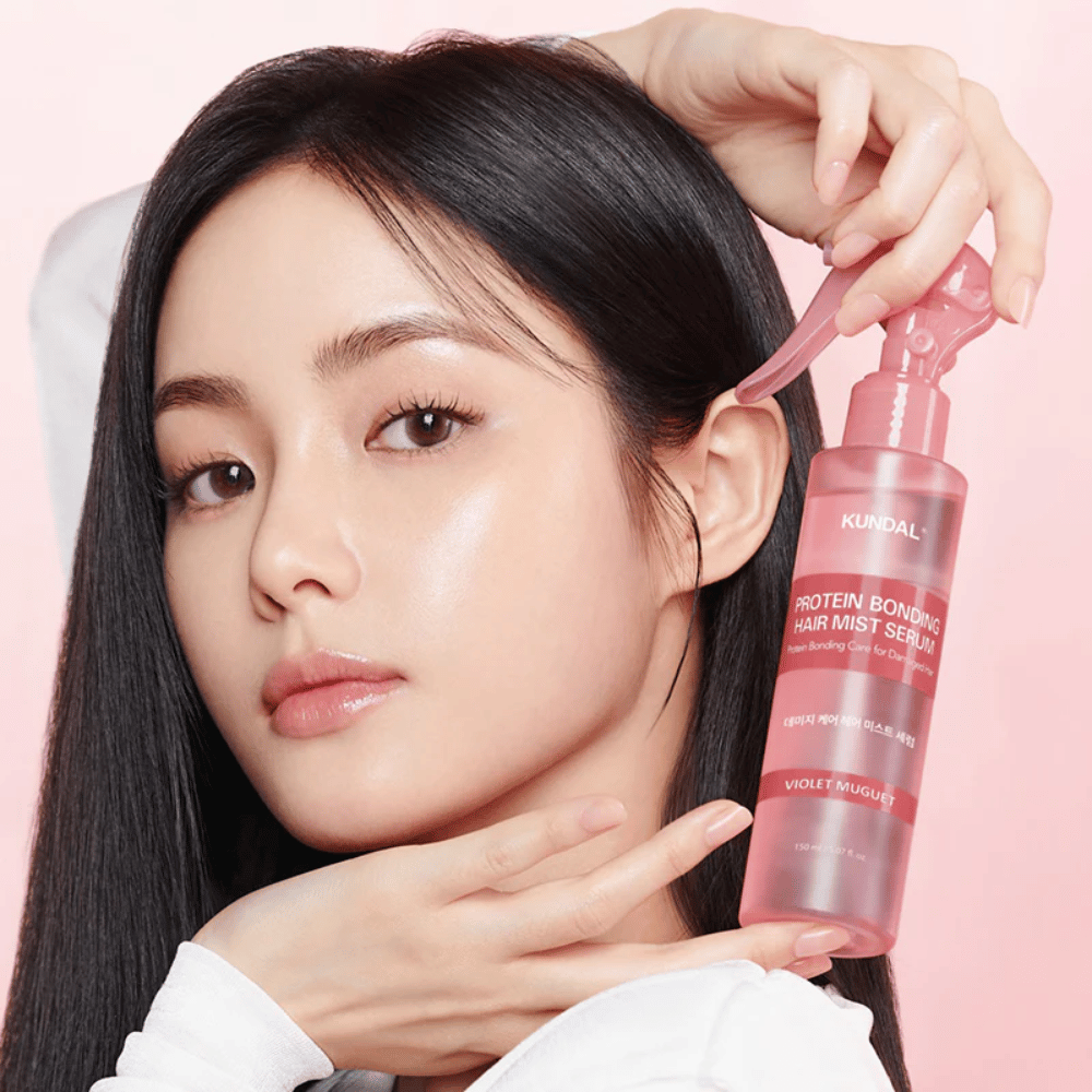 Mujer con cabello largo y negro sostiene KUNDAL Protein Bonding Hair Mist Serum Violet Muguet frente a su rostro, sobre un fondo rosa claro.