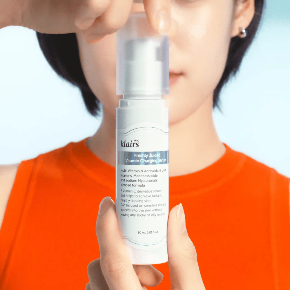 Mujer con camiseta naranja sostiene Klairs Freshly Juiced Vitamin Charging Serum frente a ella contra un fondo azul.