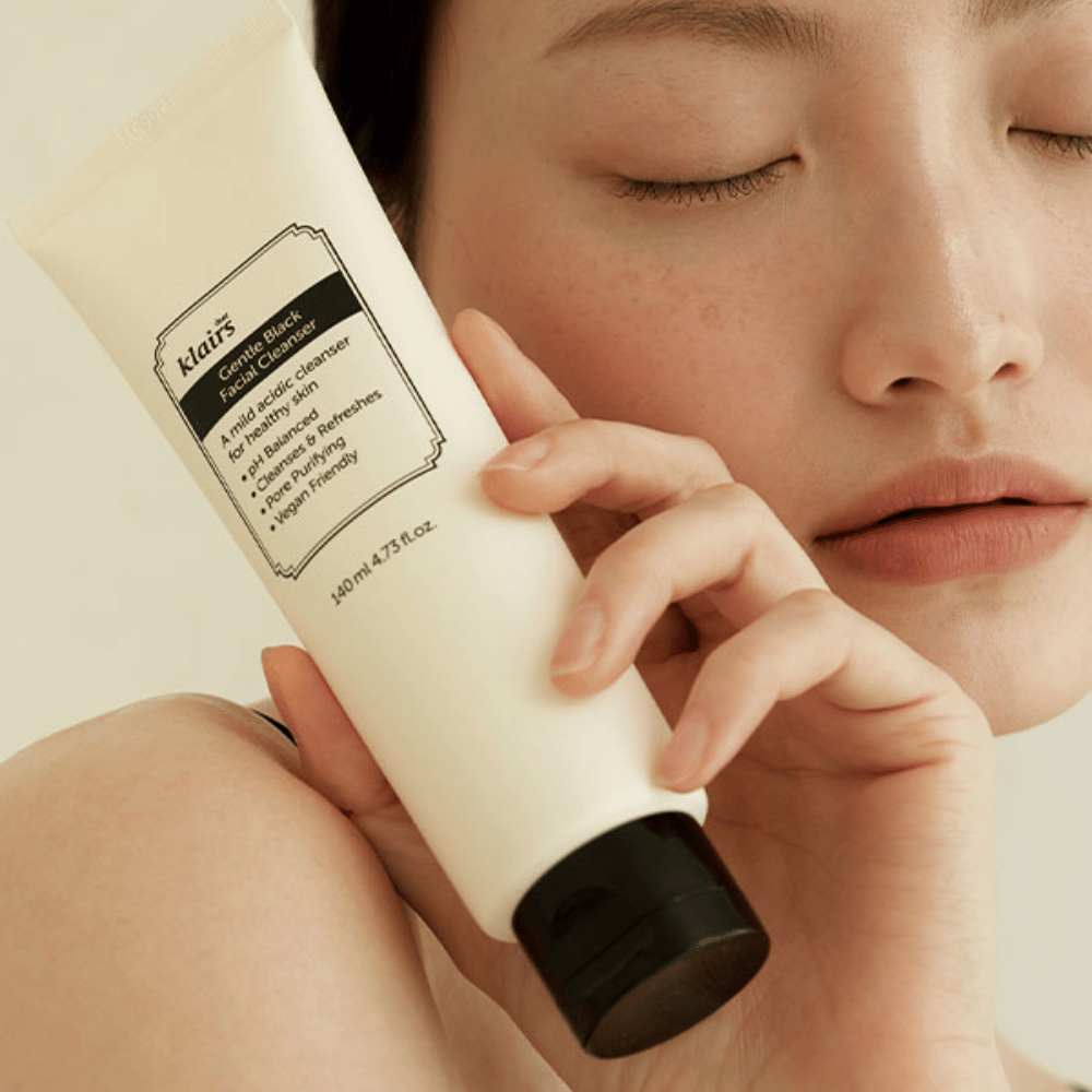 Una persona sostiene un tubo de Klairs Gentle Black Facial Cleanser contra su mejilla, ojos cerrados y tono de piel natural visible en el fondo.
