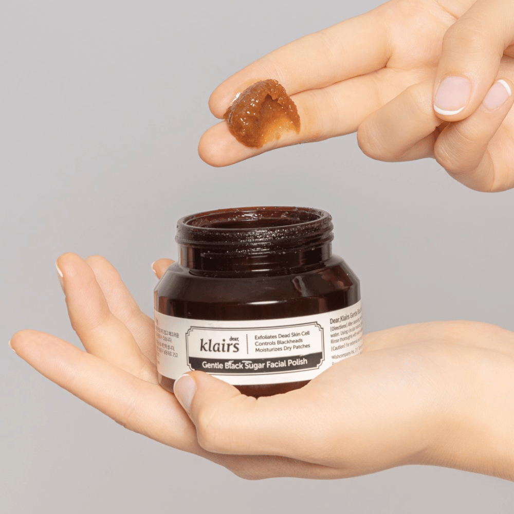 Manos sostienen un tarro abierto de Klairs Gentle Black Sugar Facial Polish y muestran el producto en un dedo, sobre un fondo gris.