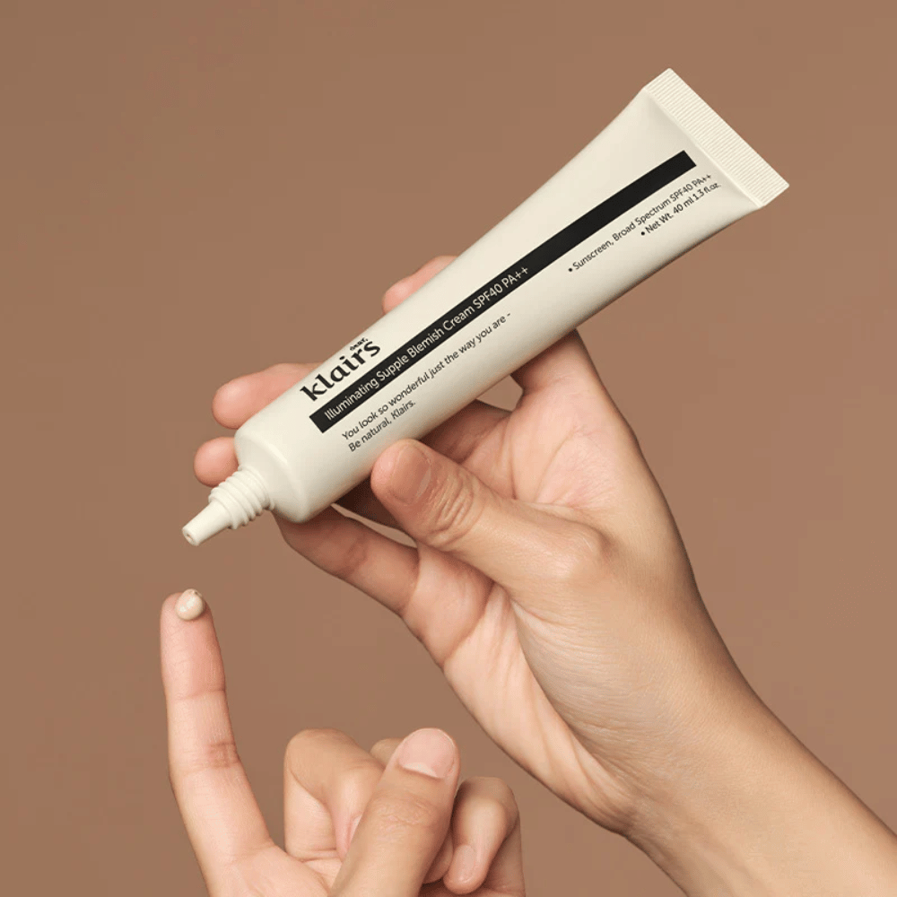 Una mano sostiene un tubo de Klairs Illuminating Supple Blemish Cream SPF40 PA++ y exprime una pequeña cantidad de crema en un dedo, sobre un fondo marrón.