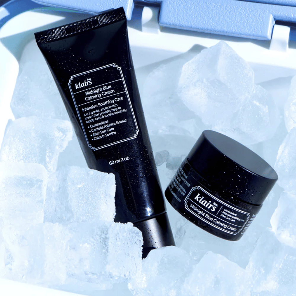 Un tubo y tarro de Klairs Midnight Blue Calming Cream sobre cubitos de hielo, con enfoque en propiedades calmantes e hidratantes, contra un fondo azul.