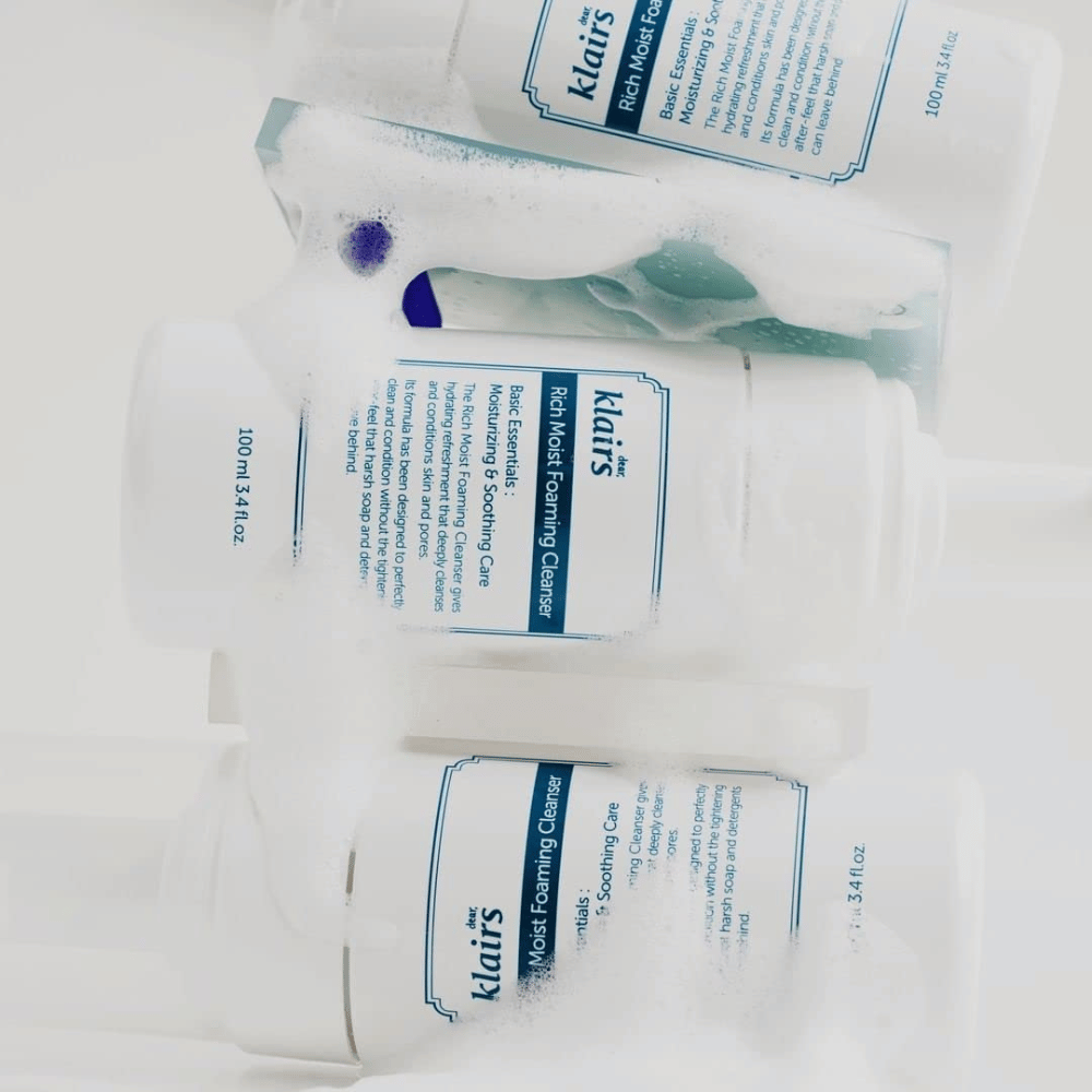 Tres frascos de Klairs Rich Moist Foaming Cleanser cubiertos de espuma y gotas de agua, colocados sobre una superficie blanca.