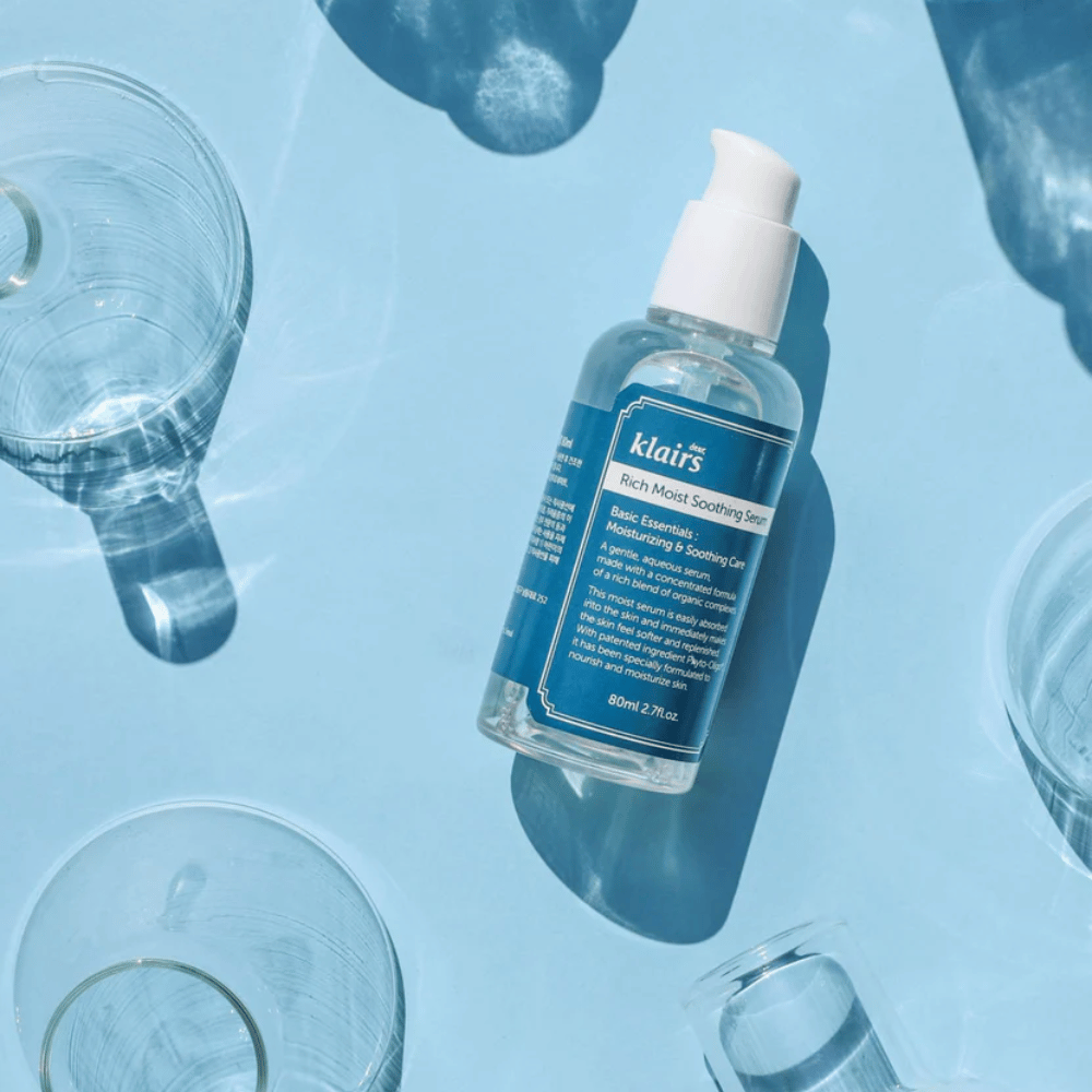 Una botella Klairs Rich Moist Soothing Serum entre vasos de agua con reflejos sobre un fondo azul.