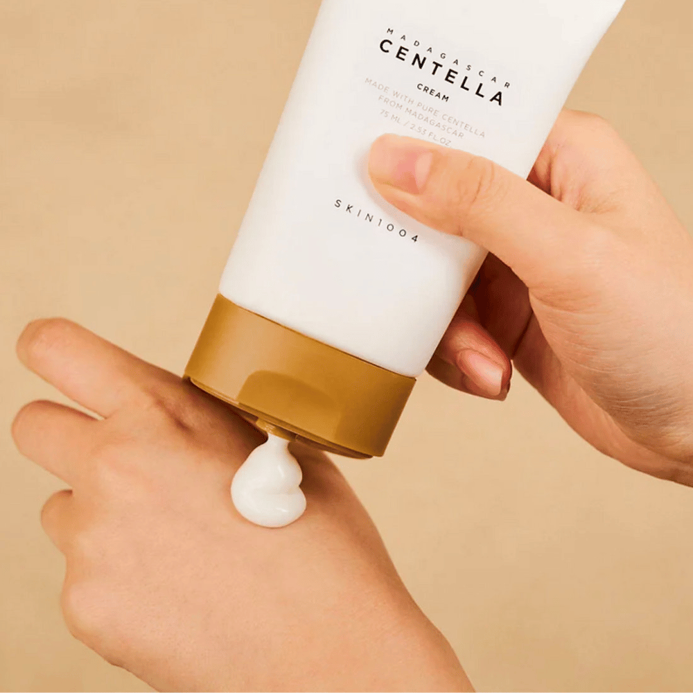 La imagen muestra a una persona sosteniendo un tubo de "Madagascar Centella Cream" de SKIN1004. Es una crema para la piel hecha de Centella pura de Madagascar, en tamaño de 75 ml / 2.53 fl.oz. La crema se aplica en la mano y está destinada a proporcionar nutrición e hidratación a la piel.