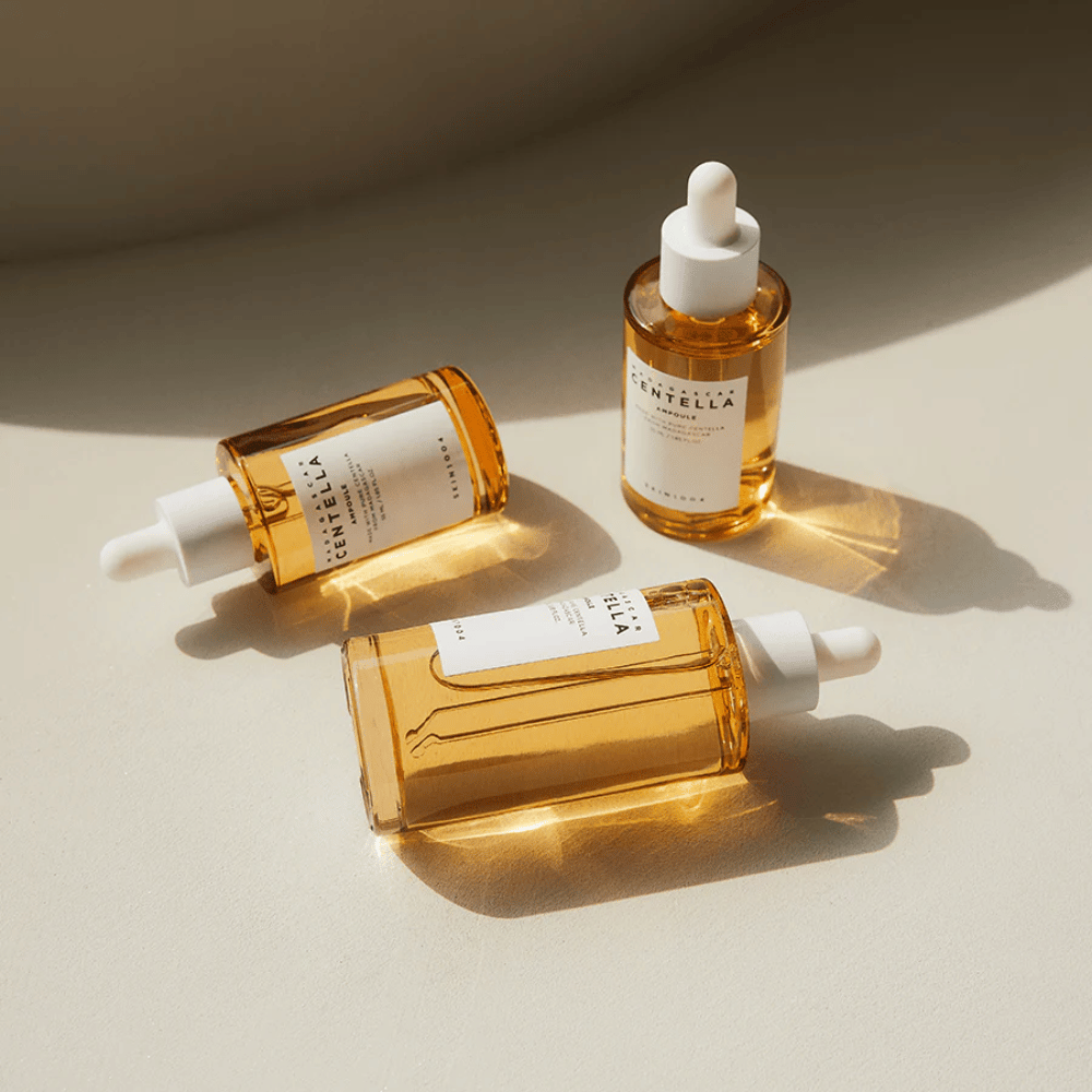 La imagen muestra tres botellas de "Madagascar Centella Asiatica 100 Ampoule". Están colocadas sobre una superficie donde la luz solar crea un hermoso reflejo a través del contenido dorado en las botellas. El producto parece ser parte de una serie de cuidado de la piel que utiliza centella asiática, un ingrediente conocido por sus propiedades calmantes. El diseño de las botellas es simple y moderno, lo que sugiere que es una serie de cuidado de la piel premium.