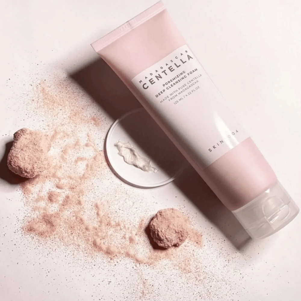 Madagascar Centella Poremizing Deep Cleansing Foam se presenta en un tubo rosa. Los tubos están sobre una superficie clara junto con polvo rosa que da una sensación de limpieza y frescura. El producto está diseñado para limpiar profundamente la piel con la ayuda de centella pura de Madagascar.