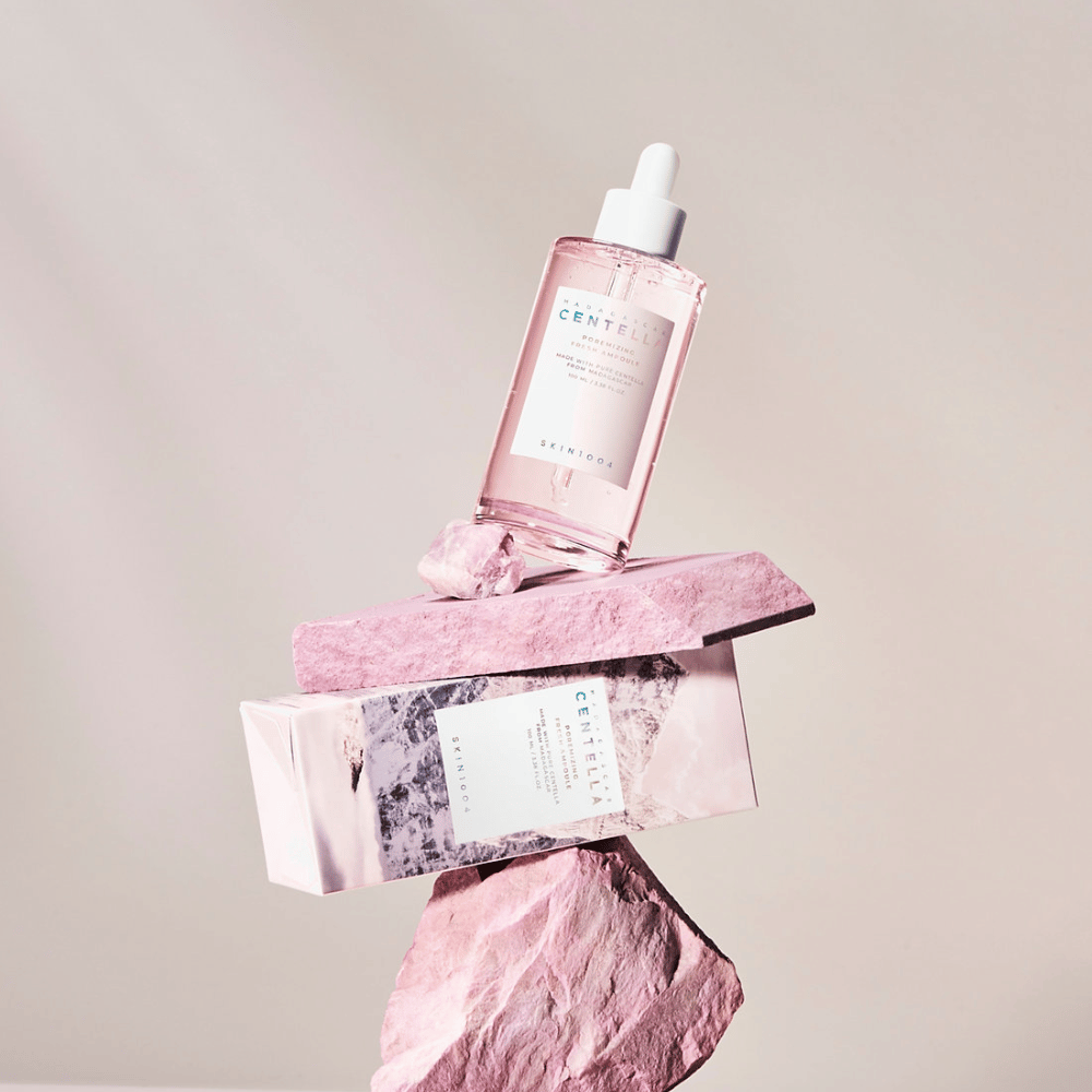 Botella transparente con ampolla de Centella rosa de SKIN1004, equilibrada sobre bloques de piedra, con un empaque de cartón debajo, sobre un fondo neutro.