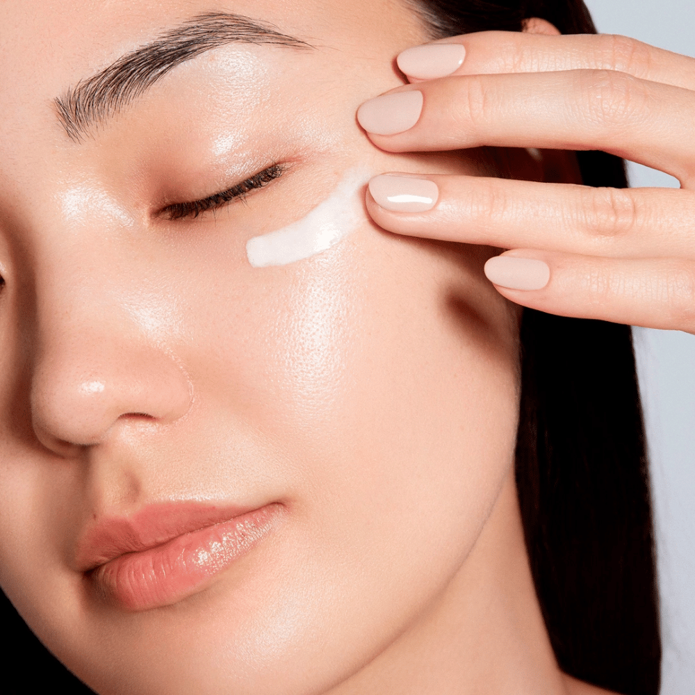 Una mujer aplica cuidadosamente una crema de ojos blanca debajo del ojo. Su piel se ve radiante e hidratada, lo que resalta el efecto de la crema de ojos. Esto destaca el producto como hidratante y adecuado para la piel sensible alrededor de los ojos.