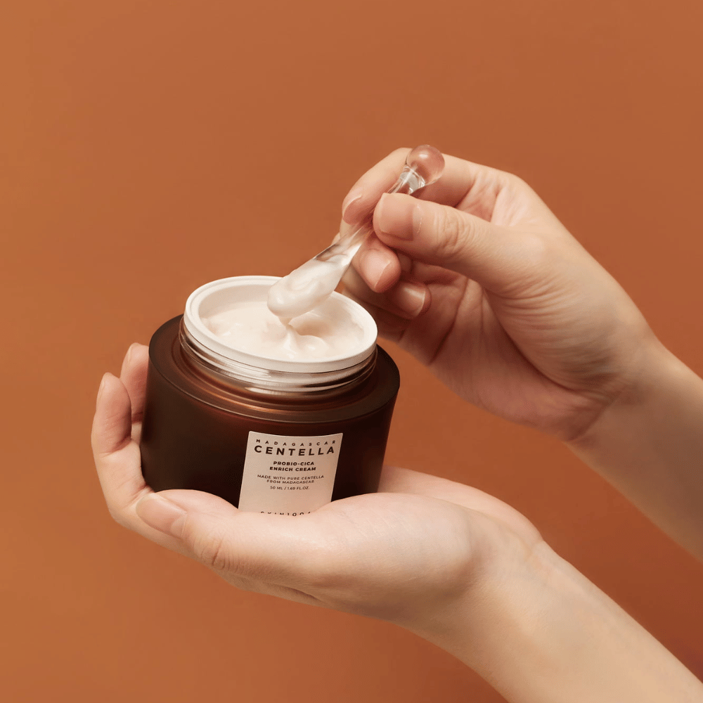 Manos sosteniendo un frasco abierto de Madagascar Centella Probio-Cica Enrich Cream y una espátula con crema contra un fondo uniforme de color terracota.