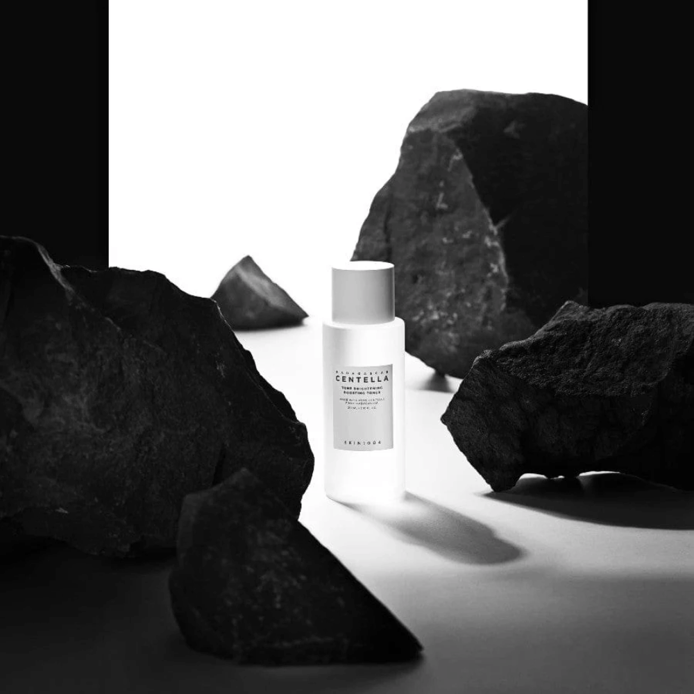 Botella blanca SKIN1004 Madagascar Centella Tone Brightening Boosting Toner  colocada dramáticamente entre grandes piedras negras sobre un fondo blanco.