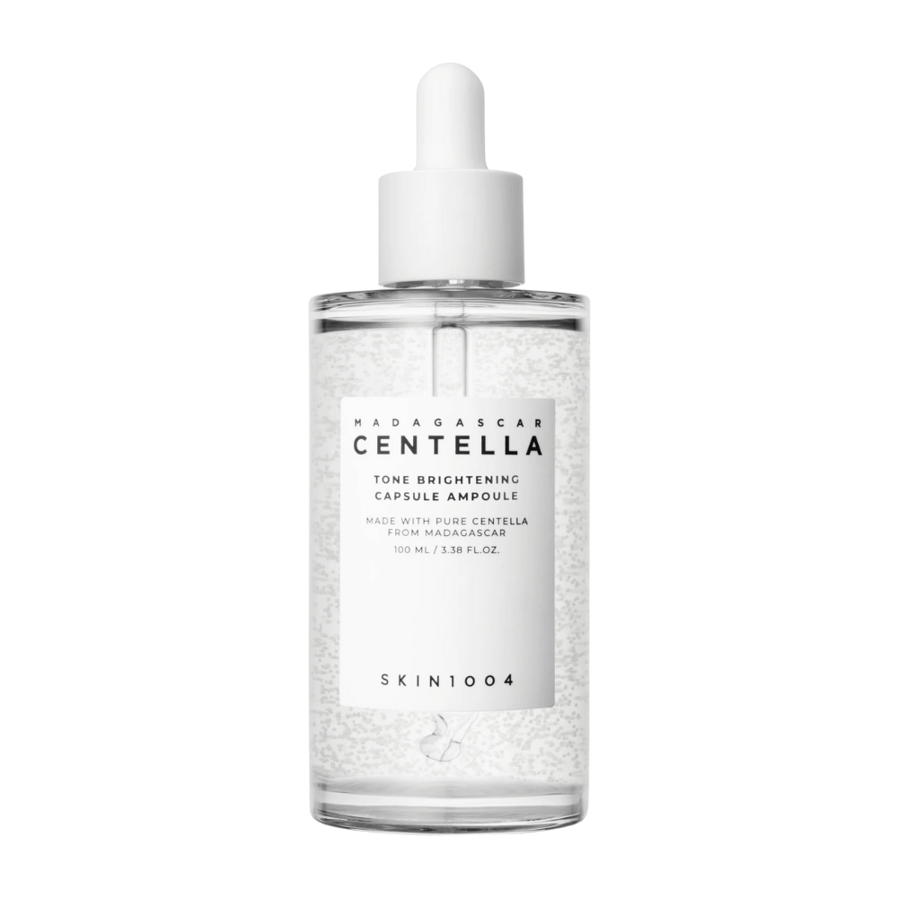 La imagen muestra una botella de "SKIN1004 Madagascar Centella Tone Brightening Capsule Ampoule". Es una botella transparente con un aplicador de gotero blanco. En la etiqueta se indica que el producto contiene ingredientes puros de centella de Madagascar. El contenido de la botella parece tener pequeñas cápsulas o partículas, lo que podría ser parte de sus propiedades aclarantes para el cuidado de la piel. La botella contiene 100 ml / 3.38 fl.oz. del producto.