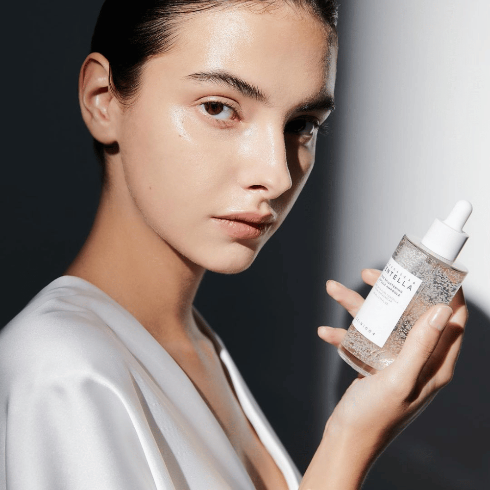 En la imagen se ve a una persona sosteniendo una botella con aplicador de gotero. La botella está etiquetada como "SKIN1004 Madagascar Centella Tone Brightening Capsule Ampoule". La persona tiene un tono de piel brillante que refleja la luz, lo que sugiere un posible efecto del producto.