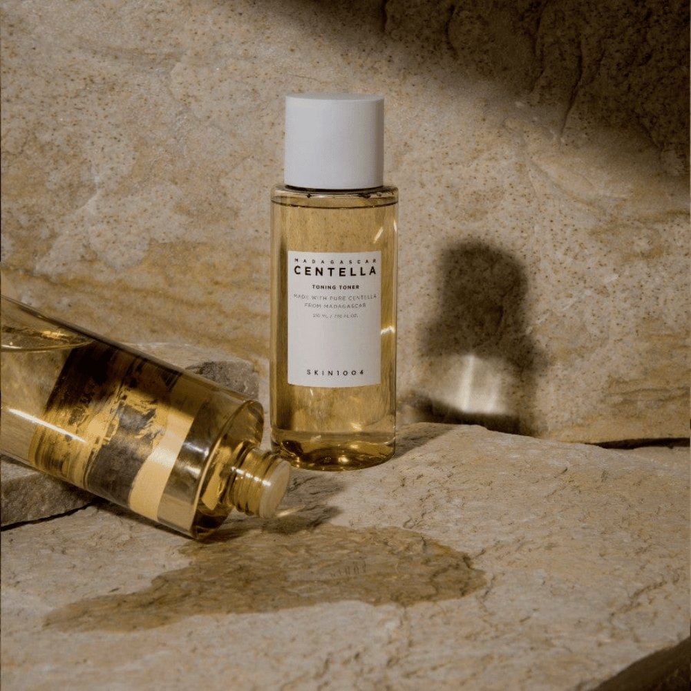 La imagen muestra una botella "Madagascar Centella Toning Toner" de SKIN1004. Es una botella transparente llena de un líquido amarillento, probablemente un tónico para el cuidado de la piel. Se indica que está hecha con centella pura de Madagascar y el contenido es de 210 ml / 7.10 fl.oz. La botella y una botella acostada están colocadas sobre una superficie rocosa, lo que proporciona una sensación natural y minimalista.