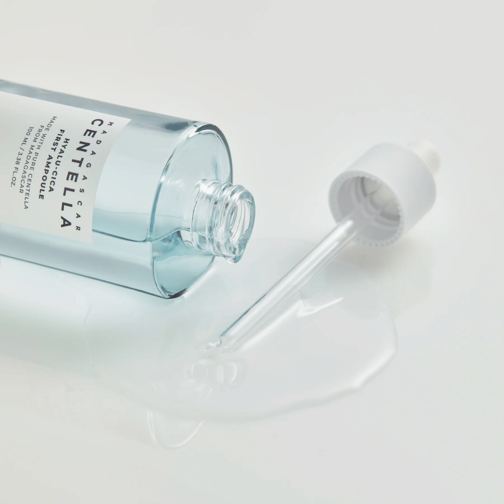 Primer plano de botella abierta Skin1004 Madagascar Centella Hyalu-Cica First Ampoule con pipeta acostada y suero esparcido en superficie blanca.