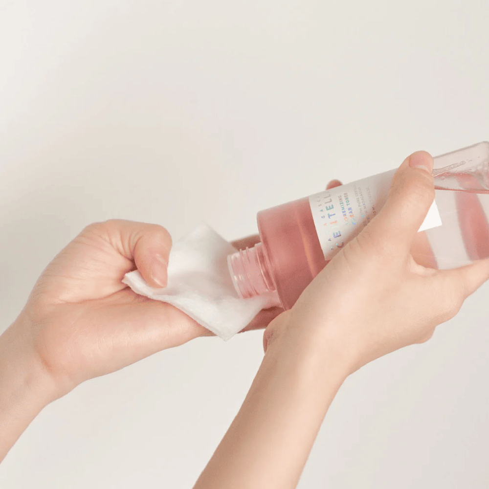 Una persona vierte Madagascar Centella Poremizing Clear Toner de una botella transparente en un disco de algodón. El producto, que tiene un tono rosa, se utiliza para refrescar y limpiar la piel. Las manos son visibles, mostrando la aplicación del tónico.
