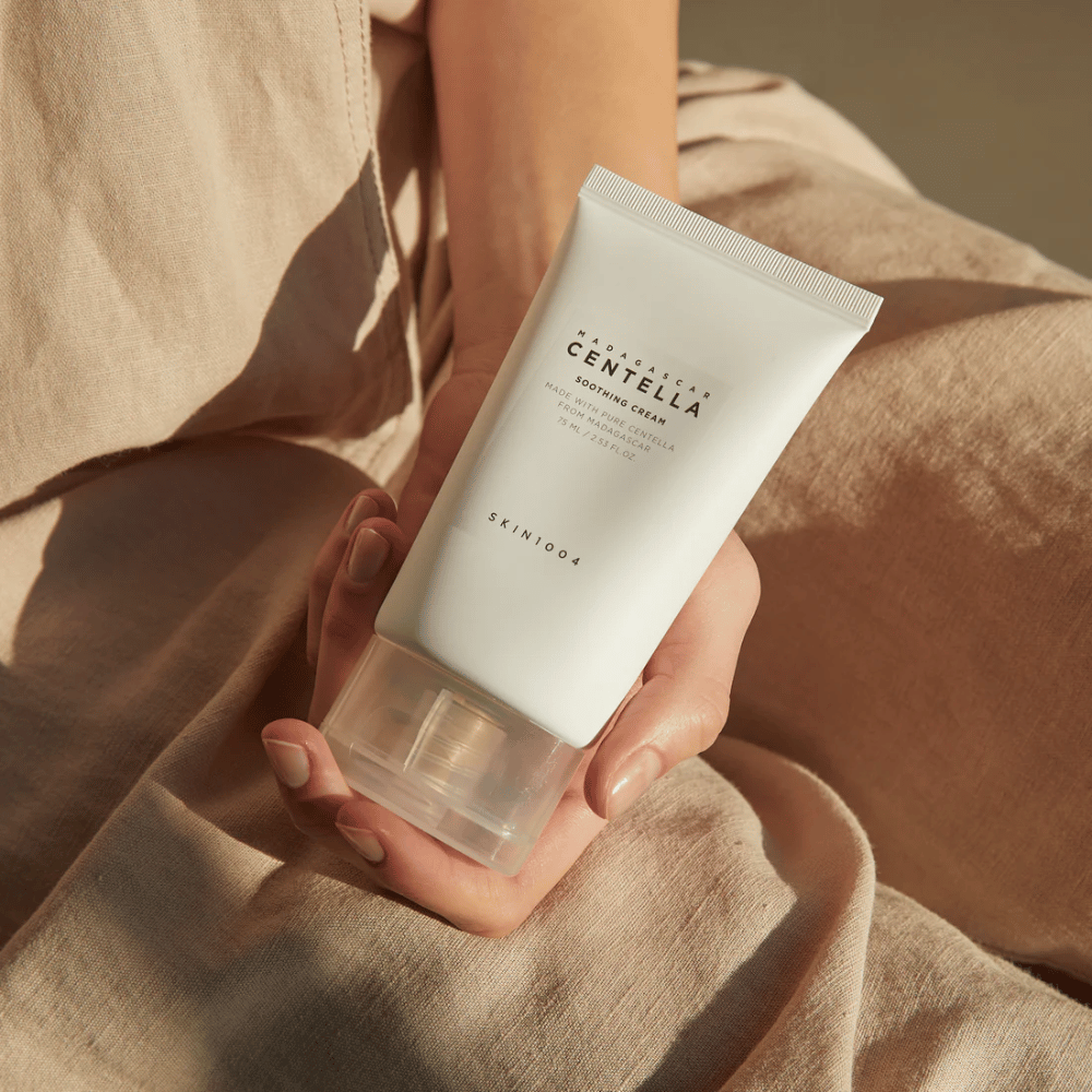 Una persona sostiene un tubo blanco de Madagascar Centella Soothing Cream de SKIN1004. El tubo tiene una tapa transparente y texto que describe el producto como calmante y fabricado con Centella pura. La persona está vestida con ropa clara.