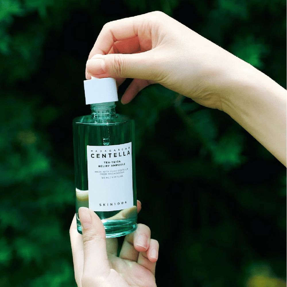 Persona abre SKIN1004 Madagascar Centella Tea-Trica Relief Ampoule – botella de vidrio verde con etiqueta blanca sostenida en las manos frente a un fondo verde.