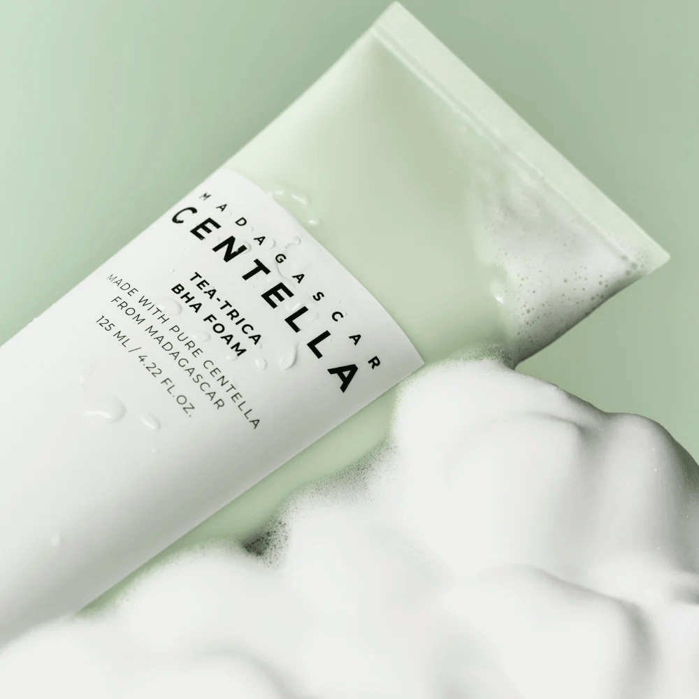 Un producto del tipo espuma limpiadora, "Madagascar Centella Tea-trica BHA Foam", es parcialmente visible con espuma alrededor. El producto tiene un diseño verde y blanco, y el texto "Madagascar Centella" y "Tea-trica BHA Foam" son claramente visibles. El volumen es de 125 ml.