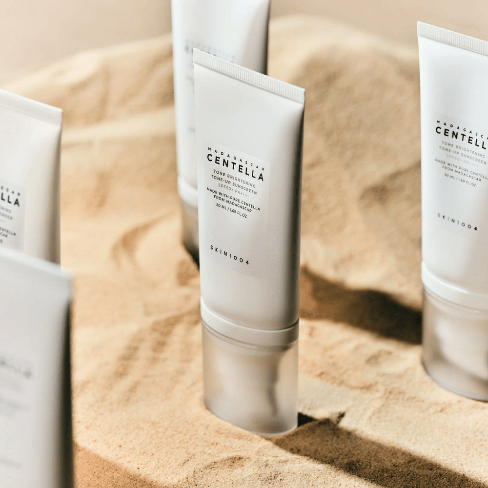 Varios tubos con Madagascar Centella Tone Brightening Tone-Up Sunscreen están en la arena. El tubo es blanco con texto negro y contiene 50 ml. El producto tiene SPF 50+ PA++++ y está fabricado con Centella pura de Madagascar.