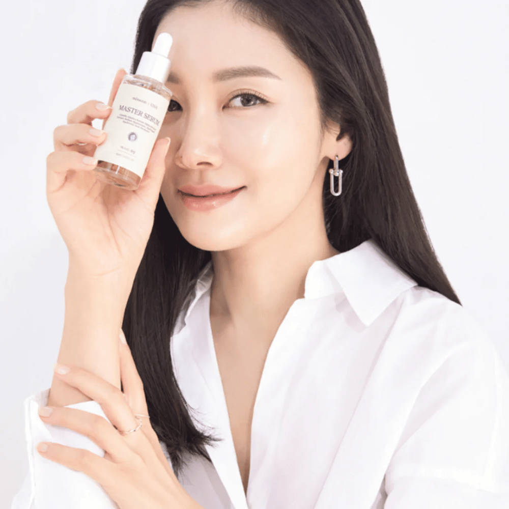 Mujer sonriente con cabello largo y oscuro y camisa blanca sostiene Mixsoon Master Serum junto a su cara. Su piel se ve radiante e hidratada.