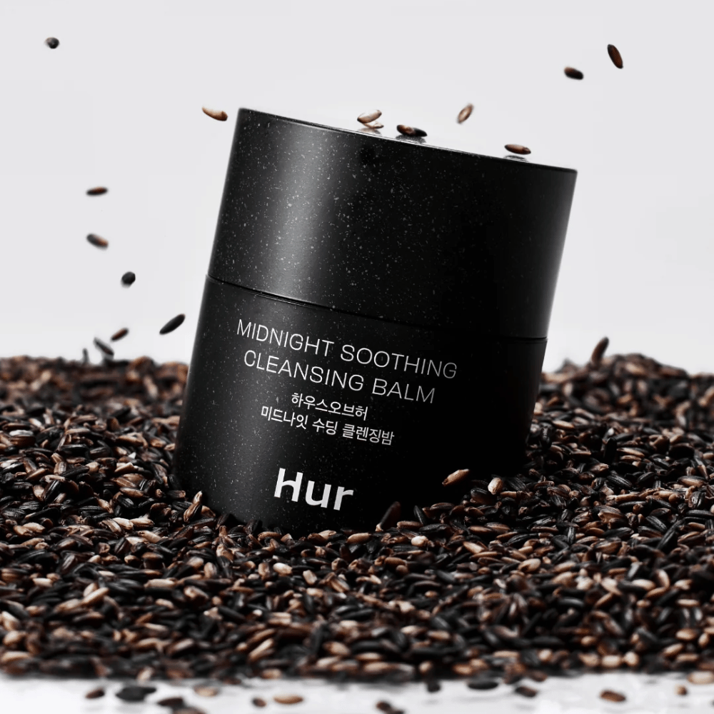 Cómo Midnight Soothing Cleansing Balm en frasco negro colocado sobre semillas negras. Bálsamo limpiador calmante que disuelve maquillaje e impurezas mientras cuida la piel.
